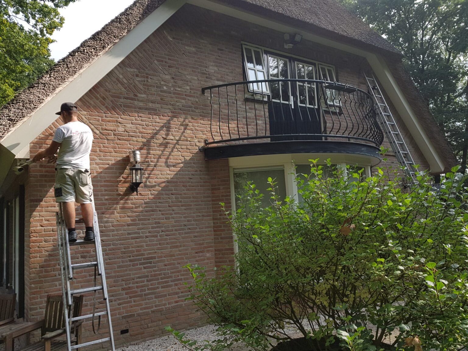 Schilder van MG Schilders op een ladder tijdens buitenschilderwerk aan een rietgedekte villa